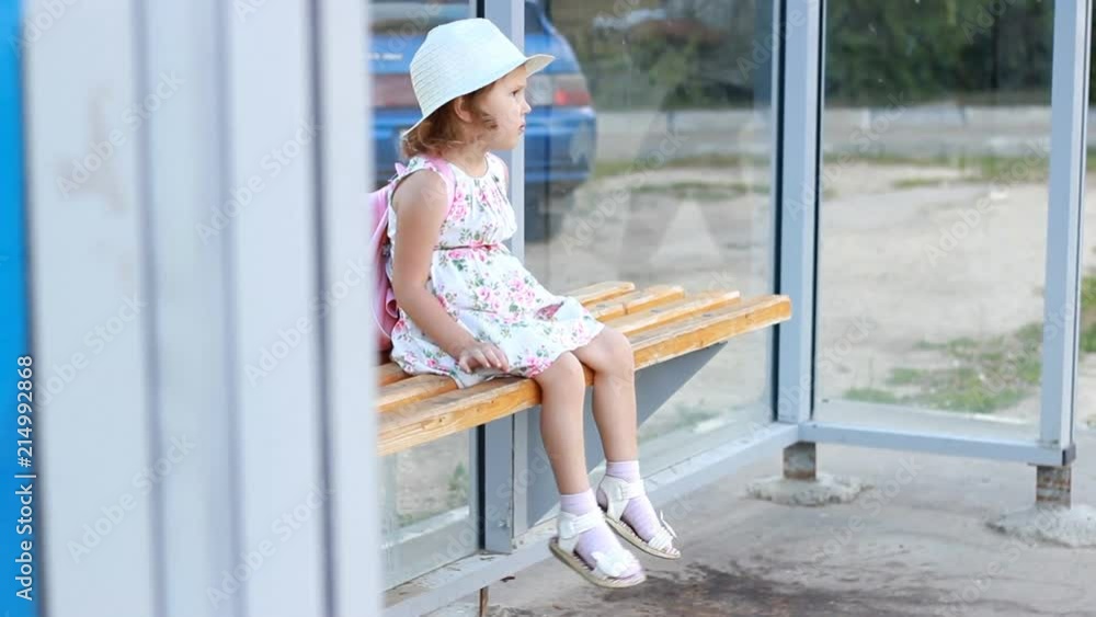 Child girl tourist in a hat and with a backpack is sitting on a bench at a bus stop, waiting for transport for a further trip Child girl tourist in a hat and with a backpack is sitting on a bench at a bus stop, waiting for transport for a further trip