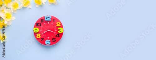 Red alarm clock with yellow daffodil flowers on a blue background flat lay top view. Place for text. Concept of Time. Long banner