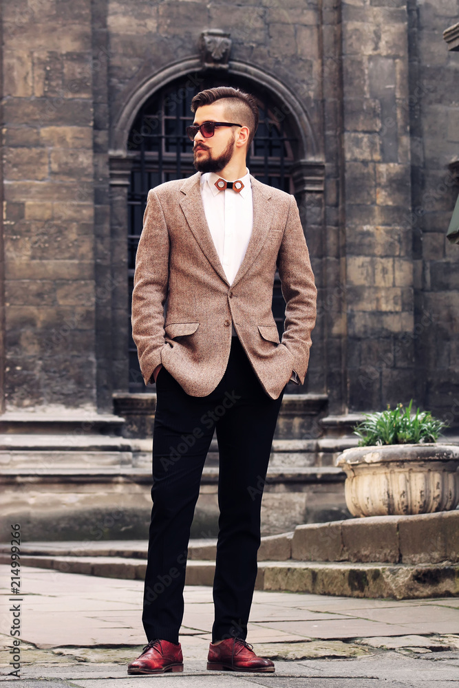 Attractive man in the street wearing british elegant suit. Young bearded businessman with modern hairstyle and sunglasses in urban background.