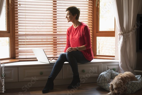 Woman looking through window while using laptop in living room photo