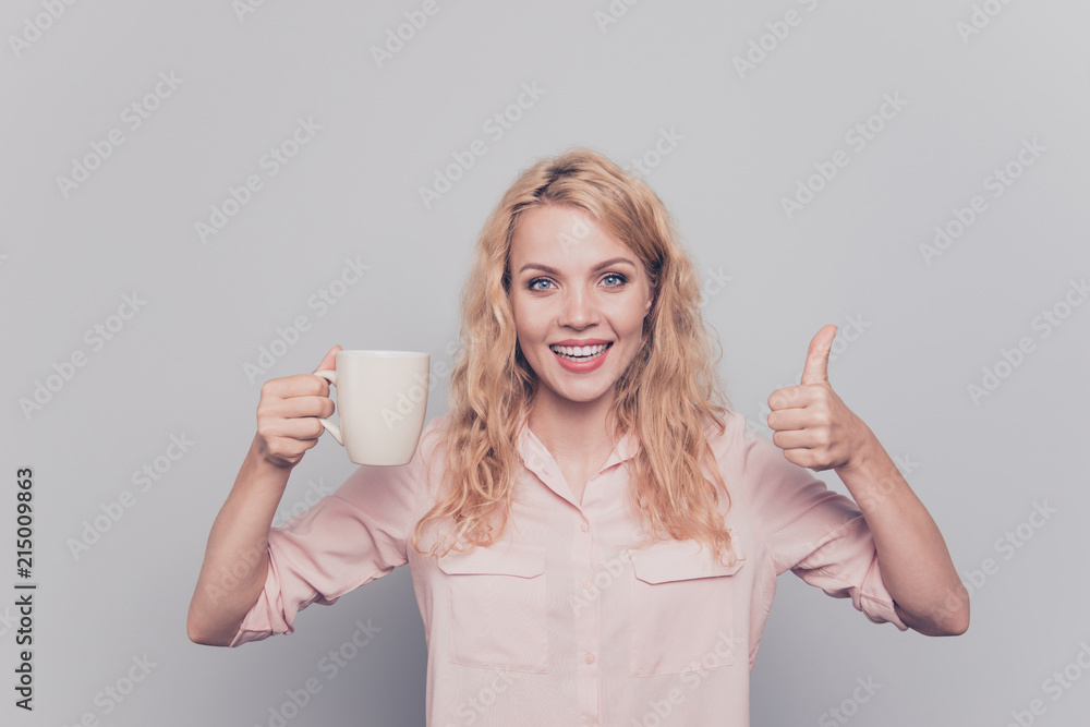 Attractive and cheerful young girl holding white mug in her hand and showing thumbs up smiling and looking at the camera isolated on gray background Attractive and cheerful young girl holding white mug in her hand and showing thumbs up smiling and looking at the camera isolated on gray background