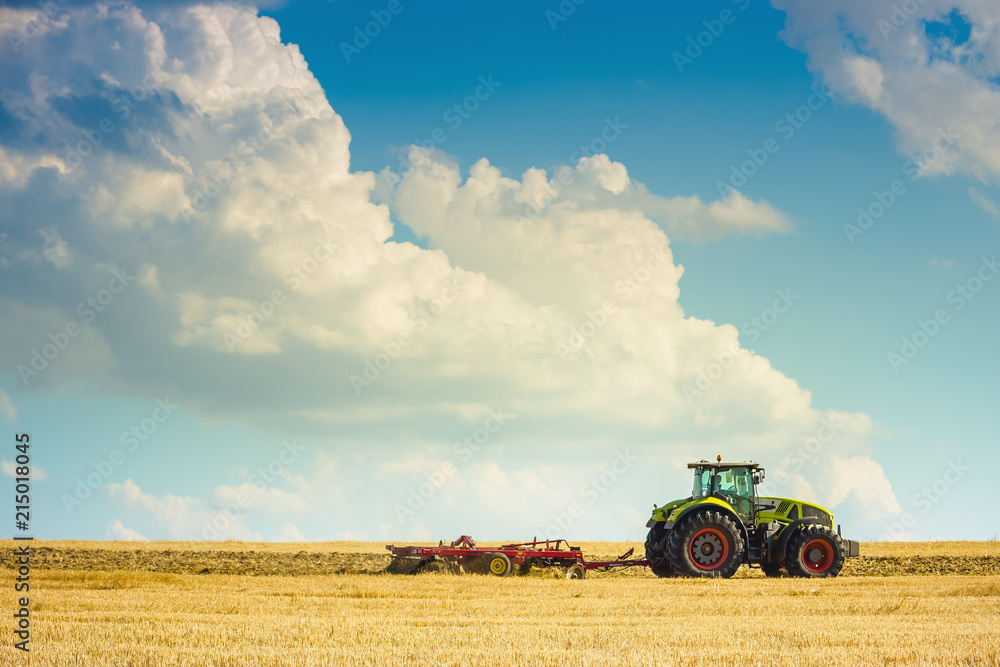 Obraz premium In the summer heat of the tractor plows the field after harvest against the bright Sunny sky with clouds.