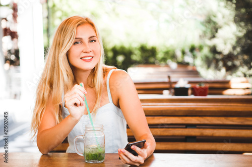 Blond girl sitting in a summer cafe with a cocktail and communicating on the Internet with a smartphone.