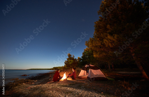 Night camping on lake shore. Active young man and woman hikers resting in front of tent at campfire under evening sky on clear blue water and green forest background. Active lifestyle concept.