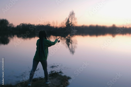 Unrecognizable woman fishing on the shore of the pond at sunset in the evening. Back view.