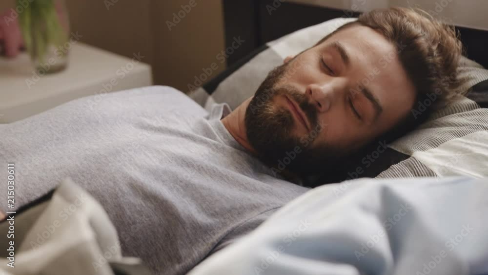 Close up of the attractive man with a beard sleeping in the bed at home, waking up and stretching while lying in the bed. Portrait shot. Indoors Close up of the attractive man with a beard sleeping in the bed at home, waking up and stretching while lying in the bed. Portrait shot. Indoors