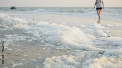 Closeup of lots of sea foam on coast during red tide algae bloom toxic beach in Naples, Florida Gulf of Mexico during sunset on sand, woman walking on coastline photo