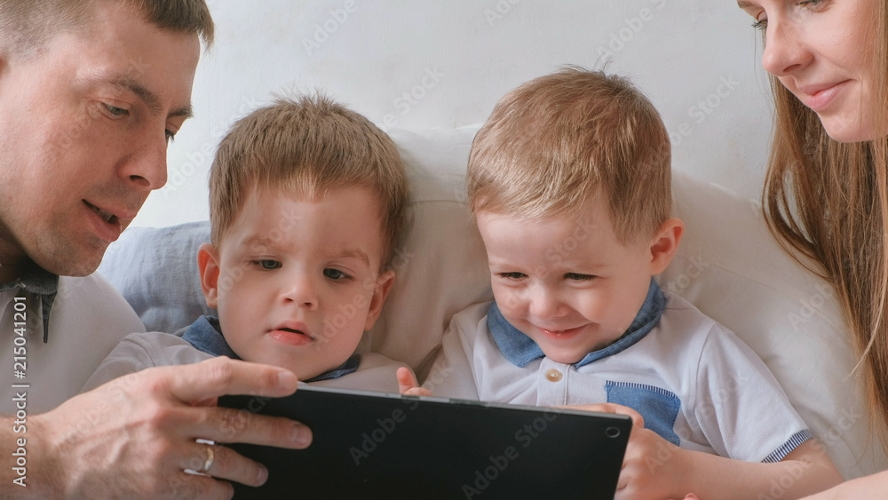 Family with tablet. Mom, dad and two sons twins toddlers looking cartoon at tablet lying on the bed.