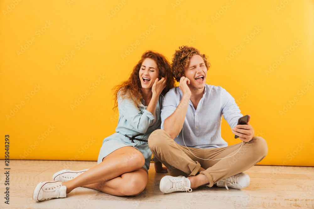 Photo of cheerful couple handsome man and curly woman 20s listening to music via earphones while sitting on floor together, isolated over yellow background Photo of cheerful couple handsome man and curly woman 20s listening to music via earphones while sitting on floor together, isolated over yellow background