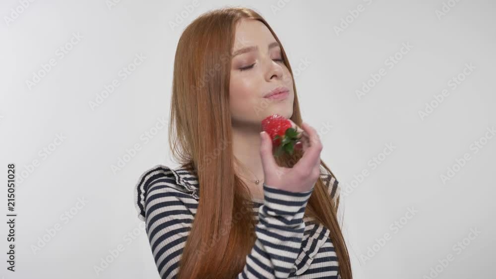 Young sweet ginger girl is biting strawberry, enjoying it, white background Young sweet ginger girl is biting strawberry, enjoying it, white background