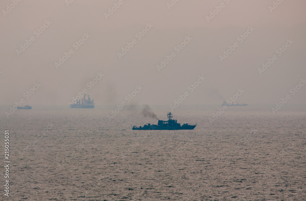 Military navy ship sailing among cargo ships on the horizon. Dark smoke coming out of a pipe