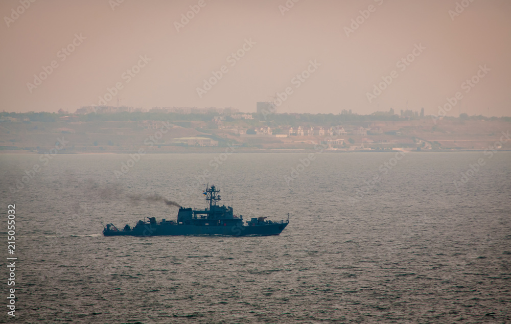 Military ship sailing in the Black sea. A shore with houses is seen at the background. Cloudy foggy weather