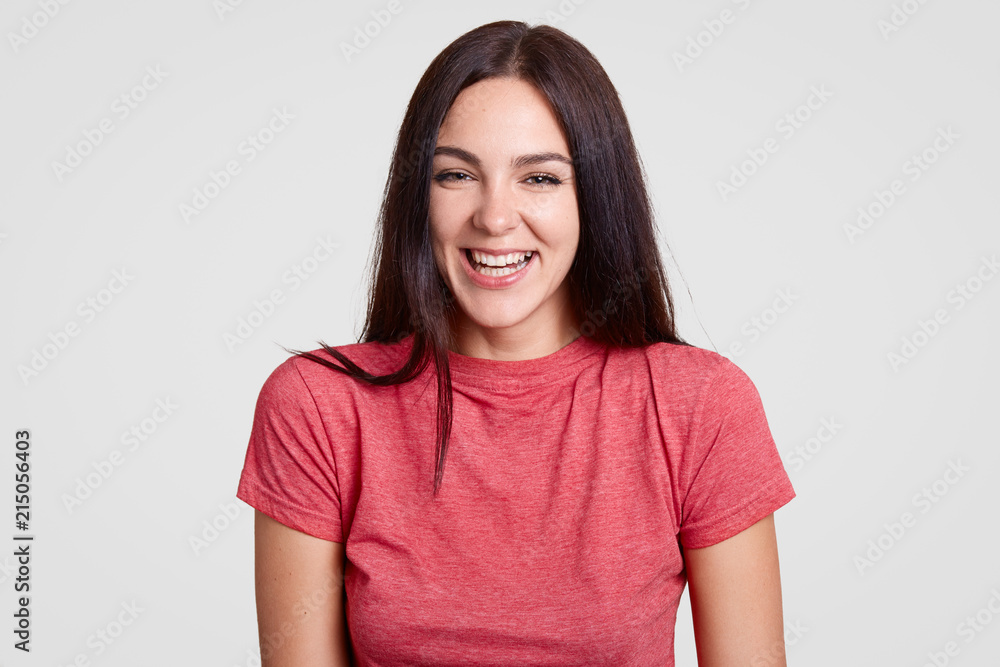 Appealing charming woman with dark hair, healthy skin, smiles joyfully, being in high spirit, wears casual pink t shirt, stands alone against white background. People, happiness and lifestyle concept Appealing charming woman with dark hair, healthy skin, smiles joyfully, being in high spirit, wears casual pink t shirt, stands alone against white background. People, happiness and lifestyle concept