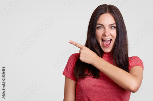 Happy brunette female opens mouth with joy, has appealing look and healthy skin, points aside at upper left corner, dressed in casual t shirt, stands against white background with free space