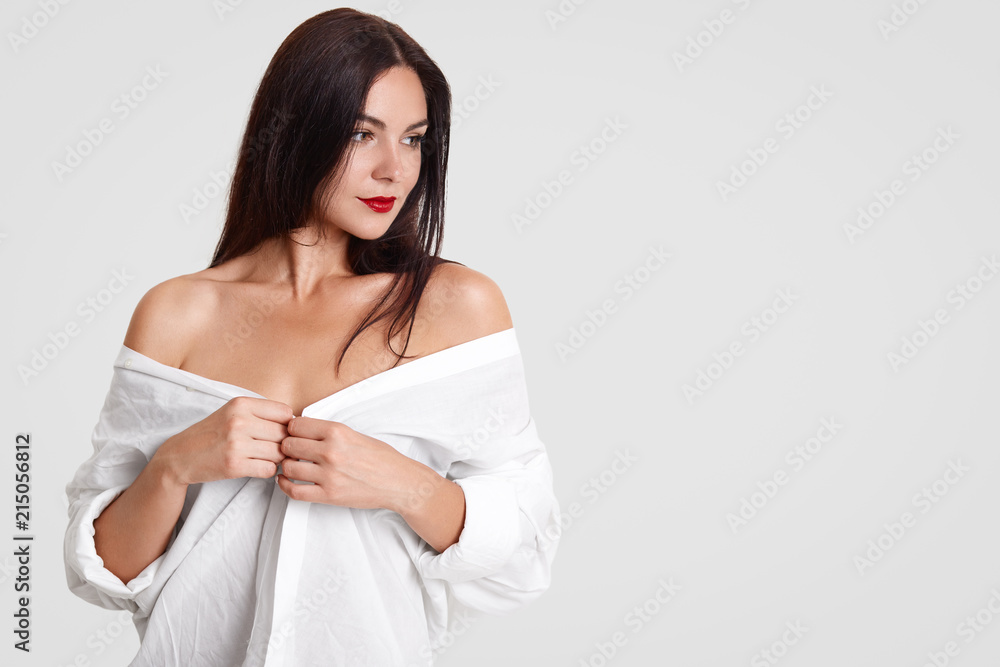 Isolated shot of pretty brunette female with sexual fit body, shows bare shoulders, wears oversized white shirt, stands against studio wall with copy space for your advertising content or text Isolated shot of pretty brunette female with sexual fit body, shows bare shoulders, wears oversized white shirt, stands against studio wall with copy space for your advertising content or text
