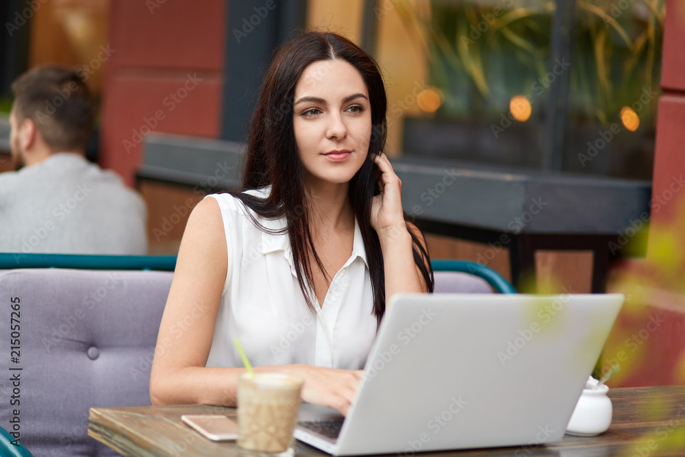 Thoughtful young female in elegant blouse, looks pensively into distnace, works on laptop computer, drinks aromatic coffee, spends free time in cafe outdoor. Woman surrounded with modern devices