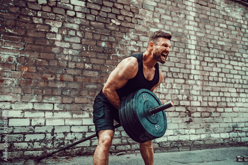 Athletic man working out with a barbell in front of brick wall. Strength and motivation. Outdoor workout. Exercise for the muscles of the back