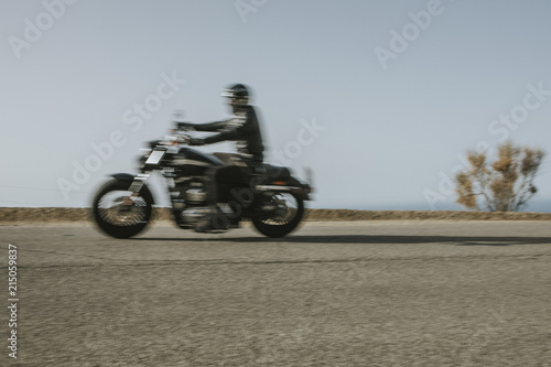 Man in black clothes riding an American motorcycle fast in a road in the mountain  with blue sky in the background.