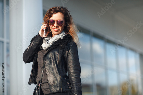 Woman in sun glasses a black leather jacket, black jeans with shopping bags talking on mobile phone in front of mirrored windows on the Street