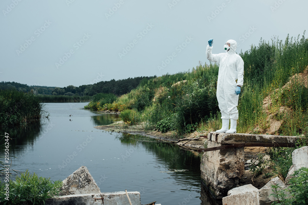 Fototapeta premium male scientist in protective mask and suit looking at sample of water in test flask outdoors
