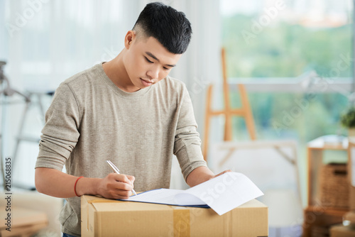 Handsome young Asian male concentrating on putting signature to document in clipboard on packed cardboard box on blurred background of art workshop 