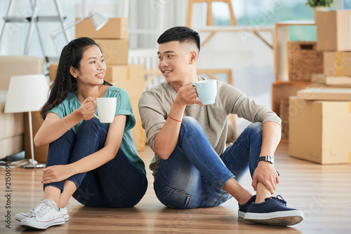 Cheerful young Asian couple sitting together with mugs of hot beverage on floor and looking at each other happily smiling on moving day with large packed cardboard boxes on blurred background .