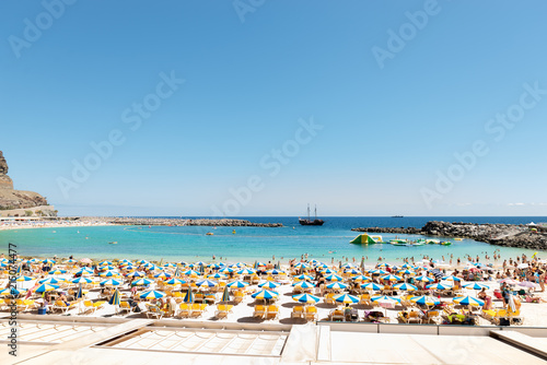 Las Americas, Canarias islands/ Spain-July 22, 2018: View of beach fool of tourists and umbrellas for sun on Canarias, view of bay with mountains. Blue Atlantic ocean and beach on Gran Canaria. © Yuliia