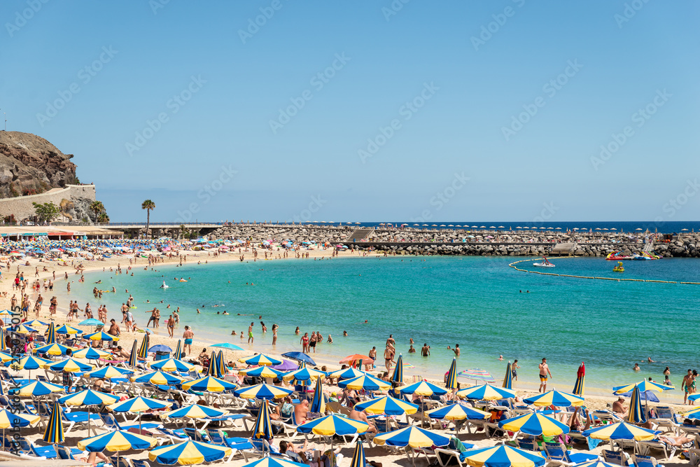 Las Americas, Canarias islands/ Spain-July 22, 2018: View of beach fool of tourists and umbrellas for sun on Canarias, view of bay with mountains. Blue Atlantic ocean and beach on Gran Canaria.