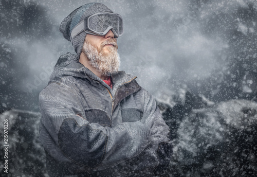 Portrait of a redhead snowboarder with a full beard in a winter hat and protective glasses dressed in a snowboarding coat posing at a studio, looking away.