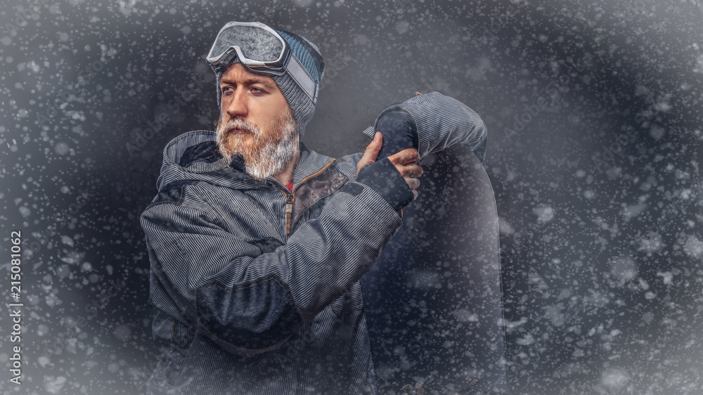 Brutal redhead snowboarder with a full beard in a winter hat and protective glasses dressed in a snowboarding coat posing with snowboard at a studio, looking away. Brutal redhead snowboarder with a full beard in a winter hat and protective glasses dressed in a snowboarding coat posing with snowboard at a studio, looking away.