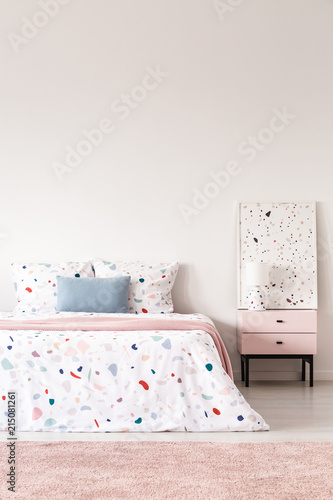 Blue pillow on patterned bed in white bedroom interior with pink carpet and poster. Real photo