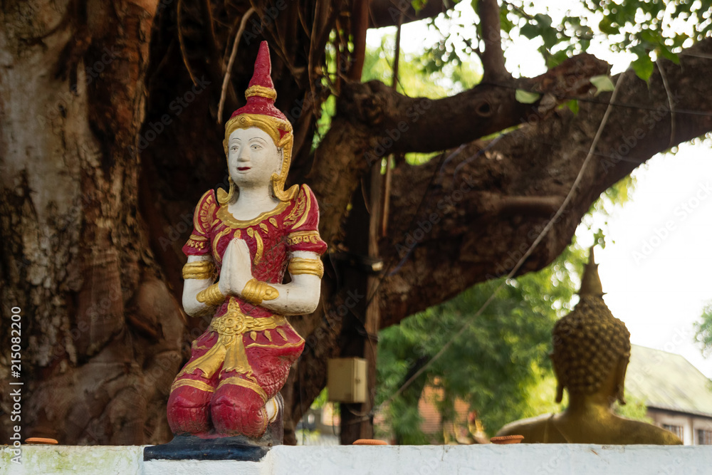 Small stone red and gold statue Buddha near to temple on a background of tree. Chiang Mai, Thailand.
