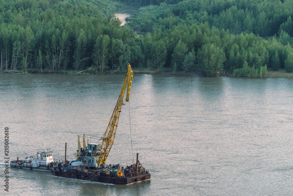 Beautiful river landscape with green shores and ship with copy space. Towboat tows barge with crane along riverbank. Dawn reflexed on calm glare water surface. Forest on background.