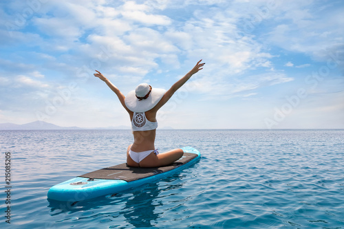Frau in weißem Bikini und mit Sonnenhut genießt ihren Urlaub auf einem Surfbrett über blauem, sommerlichen Meer