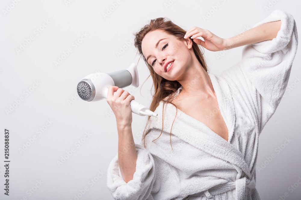 Beautiful red-haired girl in a white bathrobe bathing her hair with a hair dryer on a light background Beautiful red-haired girl in a white bathrobe bathing her hair with a hair dryer on a light background