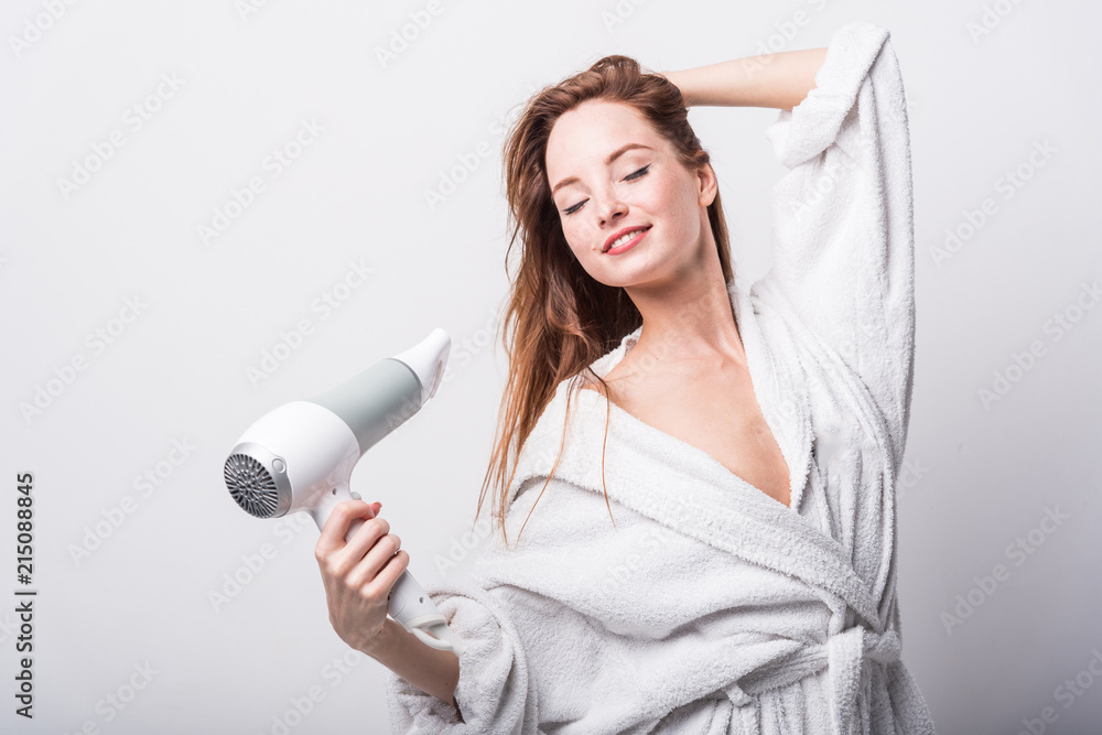 Beautiful red-haired girl in a white bathrobe bathing her hair with a hair dryer on a light background Beautiful red-haired girl in a white bathrobe bathing her hair with a hair dryer on a light background