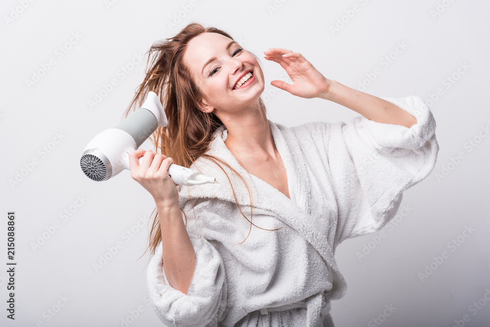 Beautiful red-haired girl in a white bathrobe bathing her hair with a hair dryer on a light background Beautiful red-haired girl in a white bathrobe bathing her hair with a hair dryer on a light background