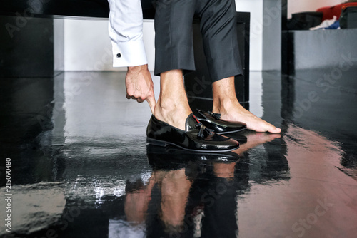 The man is wearing slip-on black glossy cut shoes, to prepare for the wedding ceremony, black wooden floor - closed up, low angle