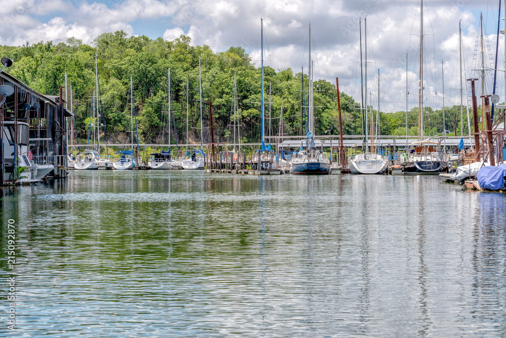 Sailboats moored in a marina under partly sunny skies against tree lined backdrop
