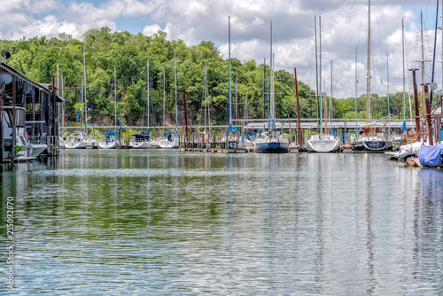 Sailboats moored in a marina under partly sunny skies against tree lined backdrop