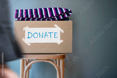 Donation Concept. Donate Box with Used Old Clothes on Chair against Wall in Public Space. blurred walking people as forground photo