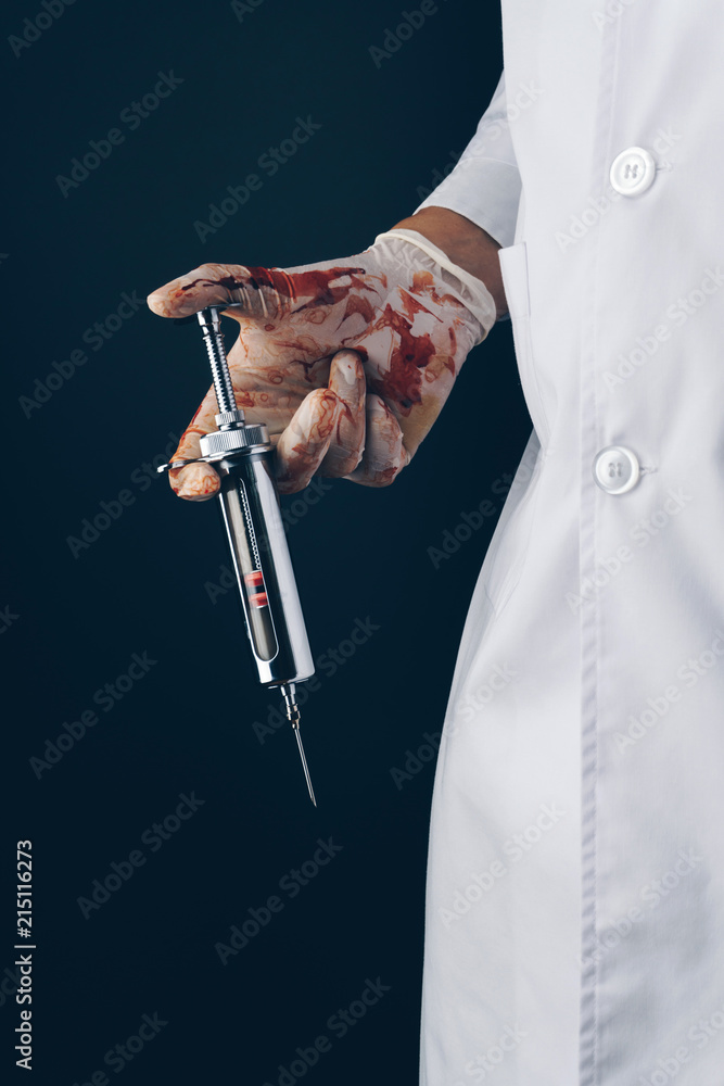 doctor wearing bloody gloves with a big stainless steel syringe on dark background