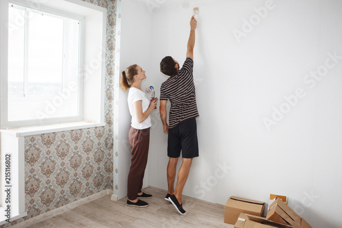 Young happy couple doing repairs in a new apartment. Painting with a roller of a white wall in a new building.