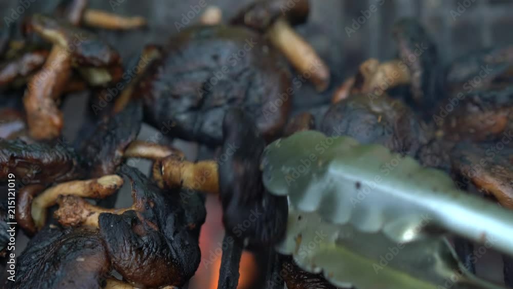 Static camera closeup of tongs turning fresh shiitake mushrooms on a hot grill with smoke and flames. Static camera closeup of tongs turning fresh shiitake mushrooms on a hot grill with smoke and flames.