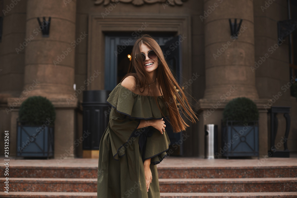 Naklejka premium Beautiful and fashionable brunette model girl with charming smile, in stylish dress with naked shoulders and in the trendy sunglasses posing outdoors in front of old building