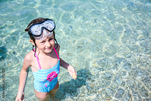 A five-year-old girl with a blurred diving mask looks directly into the camera while standing in crystal clear sea water and laughing after bathing in a bathing suit in the afternoon.