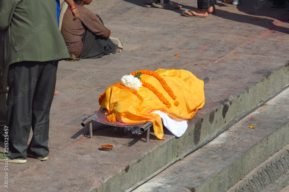 Dead body covered with white and orange textiles, waiting to be carried down the stairs to be dipped into the holy Bagmati River before cremation at the Pashupatinath Temple, Kathmandu, Nepal 