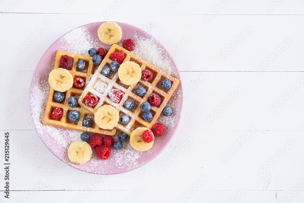 Belgian waffles with blueberries, raspberries, bananas and sugar powder on white background. Concept of tasty and healthy food. Top view.
