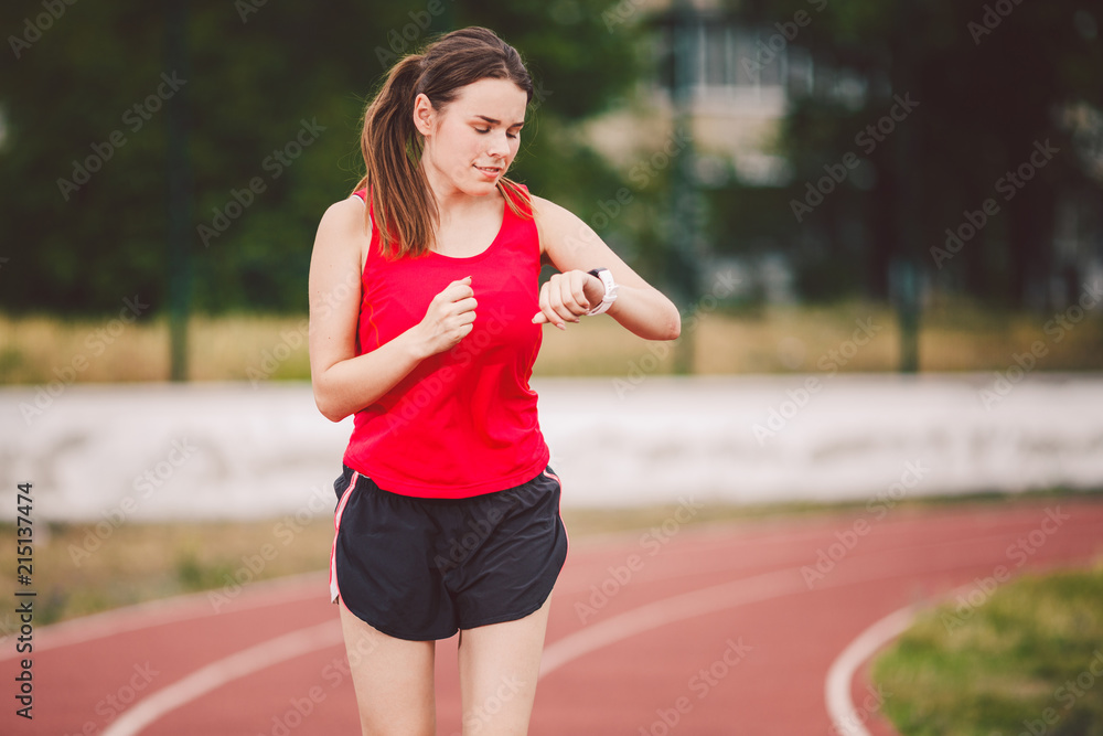 Beautiful young athlete Caucasian woman with big breast in red T-shirt and  short shorts jogging, running in the stadium. Hairstyle of a ponytail, uses  a sports watch on his arm Stock Photo