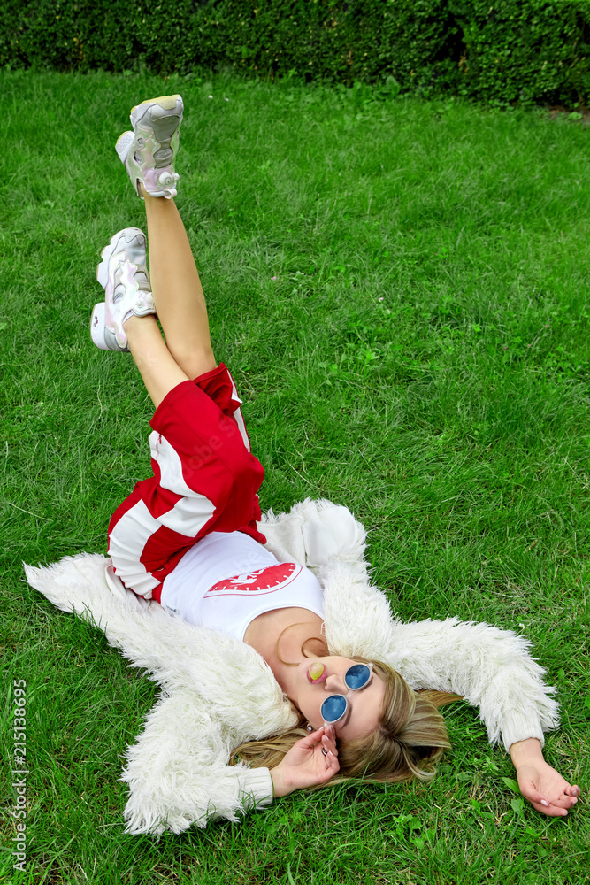 Fashion young girl laying on green grass and got over buuble of bubble gum . Stylish woman in white fur and sport red pants.Blue sunglasses.Fashion Fashion young girl laying on green grass and got over buuble of bubble gum . Stylish woman in white fur and sport red pants.Blue sunglasses.Fashion